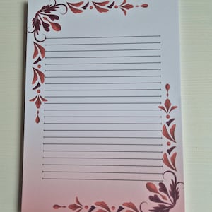 Op de afbeelding: Een gelijnd notitieblok met een roze en witte gradiënt achtergrond. Het notitieblok heeft decoratieve bordeauxrode bloemenhoekontwerpen. De lijnen zijn zwart en het papier is wit. Het notitieblok is ontworpen om op te schrijven.