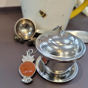 May include: A stainless steel tea infuser with a matching saucer, a tea scoop, and an owl-shaped charm with an orange stone. A white teacup with yellow handle and bee illustrations is in the background.