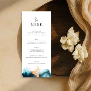 May include: A vertical menu card with the words "The Menu" in teal script. The bottom left corner features an abstract watercolour design in teal, gold, and navy. The card is on a wooden plate with white flowers and a beige fabric.