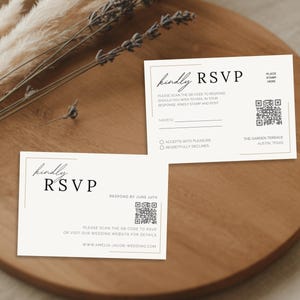 May include: Two RSVP cards with the text "kindly RSVP" in a minimalist design. The cards include QR codes and instructions for responding. The cards are on a wooden tray with dried plants.