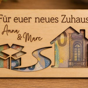 Könnte beinhalten: Eine Holz-Geschenkkarte mit dem deutschen Text "Für euer neues Zuhause" und den Namen "Anna & Marc". Die Karte zeigt Ausschnitte von einer Geschenkbox, einem gewundenen Pfad und einem Haus, wobei Währung durch die Ausschnitte sichtbar ist.