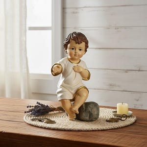 Puede incluir: Una figura de color crema de un bebé con cabello castaño, vistiendo una túnica blanca con ribetes dorados, sentado sobre una piedra gris. La figura está sobre un salvamanteles redondo y tejido con lavanda y una vela encendida.