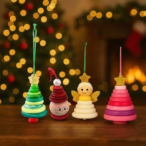 May include: Four festive Christmas tree decorations. There's a green and yellow tree, a red and pink snowman, a cream-coloured angel with gold wings, and a pink and purple tree. Each ornament is topped with a gold star and suspended by a green string.