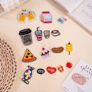 Puede incluir: Una colección de parches bordados con temas de comida y bebida. Los parches incluyen tazas de café, pizza, una magdalena, un perrito caliente y un caramelo. Otros parches representan un cartón de leche, un pez y el texto "I NEED MORE COFFEE".
