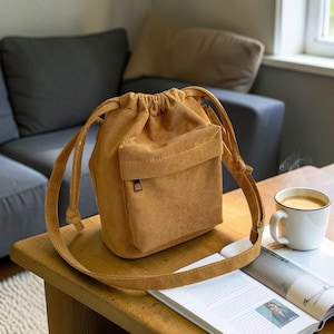 May include: A mustard-colored corduroy bucket bag with a front pocket and drawstring closure. The bag has a long, adjustable strap. The bag is sitting on a wooden table next to a cup of coffee and a magazine.