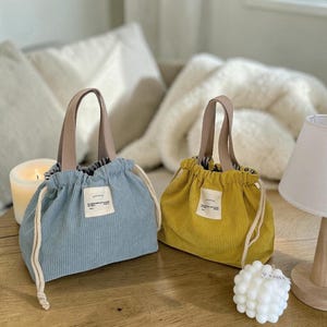 Peut inclure: Deux sacs à cordon en velours côtelé, un bleu clair et un jaune, avec des poignées et des cordons beiges. Chaque sac a une petite étiquette blanche. Les sacs sont sur une surface en bois.