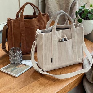 Könnte beinhalten: Zwei Cord-Tragetaschen. Eine ist warmbraun, die andere hellbeige. Beide haben obere Griffe und verstellbare Schultergurte. Jede Tasche hat eine Vordertasche und ein kleines Etikett. Die Taschen stehen auf einer Holzoberfläche.