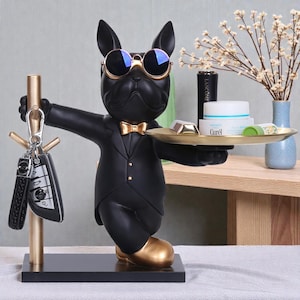 Op de afbeelding: Een zwart bulldogbeeldje met een zonnebril, een gouden vlinderdas en een smoking. Het beeldje houdt een gouden dienblad met items en een gouden sleutelhouder met sleutels vast. De bulldog heeft gouden schoenen.