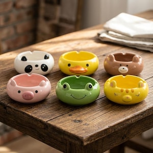 Può includere: Una collezione di sei posacenere in ceramica con diversi design di animali, tra cui un panda, un'anatra, un orso, un maiale, una rana e un gatto. I posacenere dai colori vivaci sono su un tavolo di legno rustico. Ogni posacenere ha una forma circolare con un piccolo bordo.