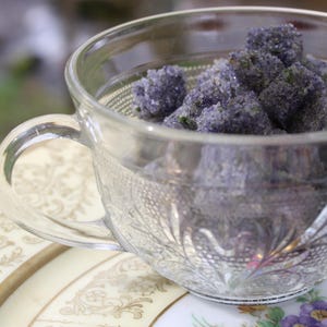 May include: A clear glass teacup filled with lavender-colored sugar cubes. The teacup has a handle and is resting on a decorative plate with floral patterns. The sugar cubes have a textured, crystalline appearance.