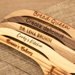 Könnte beinhalten: Eine Auswahl an Brotschneidemessern aus Holz mit Gravuren. Die Texte lauten "Bread Queen", "Create delicious", "DR. LENA BRUHIN", "Carly's Kitchen" und "Bennie's Bakery". Die Messer sind aus verschiedenen Holzarten gefertigt und haben einen Schneidedraht.
