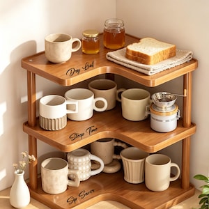 Op de afbeelding: Een houten hoekplank met drie niveaus en een lichtbruine houtnerfafwerking. De planken zijn voorzien van zwarte tekstlabels met Dry Leaf, Steep Time en Sip Savor. Het rek bevat diverse keramische mokken, glazen potten en een snee brood op een doek.