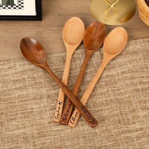 May include: Four wooden spoons with engraved names: Sophia, Olivia, Ethan, and SMITH. The spoons vary in color from light to dark brown. The spoons are arranged on a tan fabric surface.