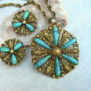 May include: A gold tone necklace and earrings set with turquoise and purple stones. The pendant is a large round design with a central pearl and turquoise stones radiating out. The earrings are smaller versions of the pendant.