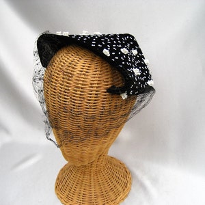 May include: A black velvet hat with white polka dots and a black net veil. The hat is perched on a wicker hat form.