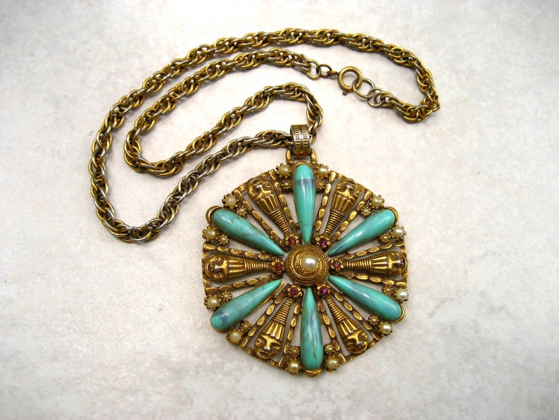 May include: A gold tone necklace with a large pendant featuring a turquoise and red stone design. The pendant is shaped like a flower with a central pearl.