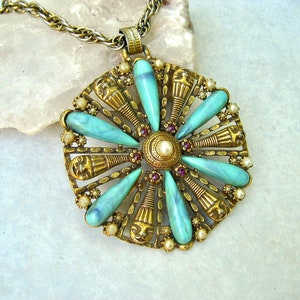 May include: A gold tone necklace with a large pendant featuring a turquoise and gold design. The pendant is shaped like a flower with a central pearl and red rhinestones.