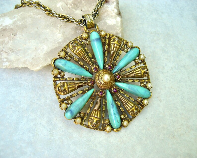 May include: A gold tone necklace with a large pendant featuring a turquoise and gold design. The pendant is shaped like a flower with a central pearl and red rhinestones.