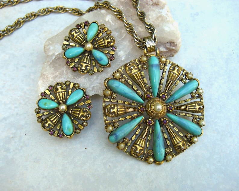 May include: A gold tone necklace and earrings set with turquoise and purple stones. The pendant is a large round design with a central pearl and turquoise stones radiating out. The earrings are smaller versions of the pendant.