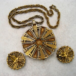 May include: A gold tone necklace with a large, round pendant featuring a sunburst design with green and gold accents. The pendant is attached to a gold tone chain. The necklace comes with a pair of matching earrings.