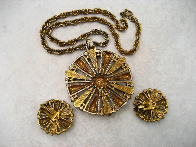 May include: A gold tone necklace with a large, round pendant featuring a sunburst design with green and gold accents. The pendant is attached to a gold tone chain. The necklace comes with a pair of matching earrings.