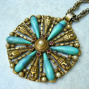 May include: A gold tone pendant necklace with a turquoise and pearl centrepiece. The pendant features a circular design with a central pearl surrounded by turquoise stones and gold accents. The necklace is attached to a silver chain.