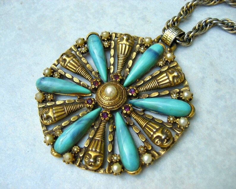 May include: A gold tone pendant necklace with a turquoise and pearl centrepiece. The pendant features a circular design with a central pearl surrounded by turquoise stones and gold accents. The necklace is attached to a silver chain.