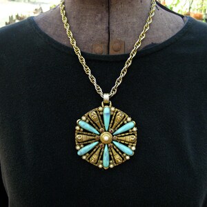 May include: A gold chain necklace with a large pendant featuring a turquoise and gold design. The pendant is circular and has a flower-like shape with eight points. Each point has a turquoise stone and a gold accent. The centre of the pendant has a pearl and red stones.