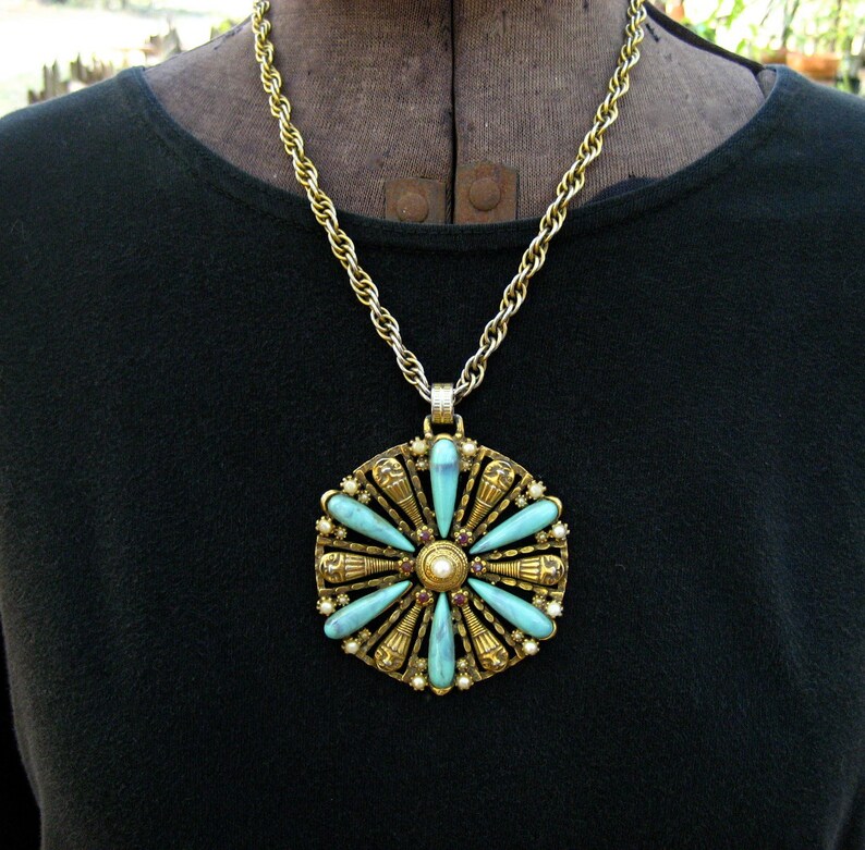 May include: A gold chain necklace with a large pendant featuring a turquoise and gold design. The pendant is circular and has a flower-like shape with eight points. Each point has a turquoise stone and a gold accent. The centre of the pendant has a pearl and red stones.