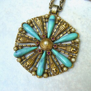 May include: A gold tone pendant necklace with a large, ornate, turquoise and gold flower-shaped pendant. The pendant features a central circle with a pearl and is surrounded by eight turquoise teardrop-shaped stones.