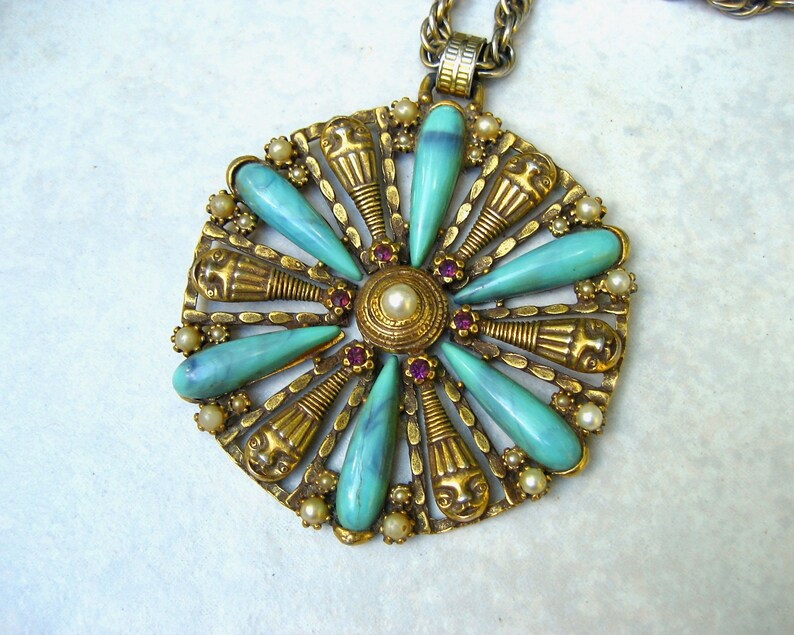 May include: A gold tone pendant necklace with a large, ornate, turquoise and gold flower-shaped pendant. The pendant features a central circle with a pearl and is surrounded by eight turquoise teardrop-shaped stones.