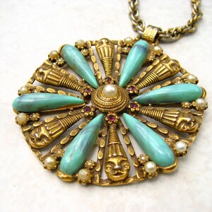 May include: A gold tone pendant necklace with a large, circular pendant featuring a turquoise and gold design. The pendant is adorned with small, white pearls and red rhinestones.