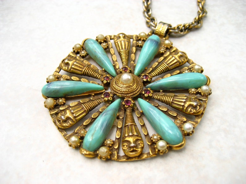May include: A gold tone pendant necklace with a large, circular pendant featuring a turquoise and gold design. The pendant is adorned with small, white pearls and red rhinestones.