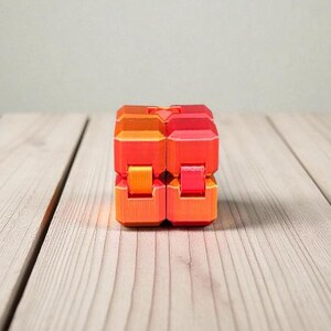 K&ouml;nnte beinhalten: Ein zweiteiliges Fidget-Spielzeug in Orange- und Rott&ouml;nen liegt auf einer hellen Holzoberfl&auml;che. Das Spielzeug besteht aus mehreren rechteckigen Bl&ouml;cken, die miteinander verbunden zu sein scheinen. Der Hintergrund ist einfarbig hellgrau.