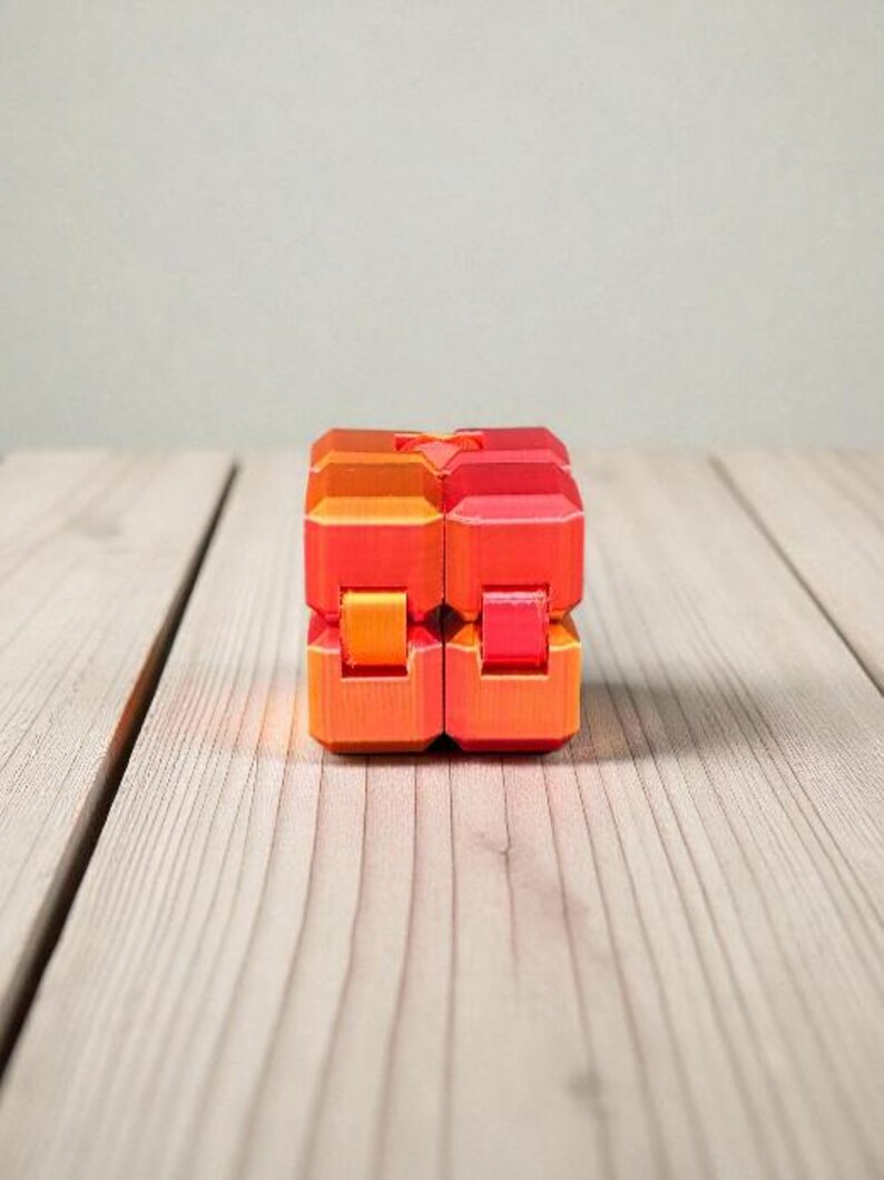 K&ouml;nnte beinhalten: Ein zweiteiliges Fidget-Spielzeug in Orange- und Rott&ouml;nen liegt auf einer hellen Holzoberfl&auml;che. Das Spielzeug besteht aus mehreren rechteckigen Bl&ouml;cken, die miteinander verbunden zu sein scheinen. Der Hintergrund ist einfarbig hellgrau.