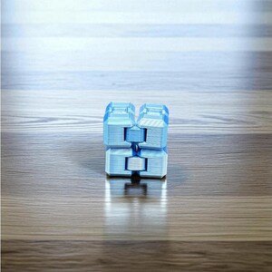 K&ouml;nnte beinhalten: Ein hellblaues Fidget-Cube-Spielzeug mit geometrischem Design. Der W&uuml;rfel besteht aus mehreren verbundenen Bl&ouml;cken, die es erm&ouml;glichen, ihn zu manipulieren und zu bewegen. Das Spielzeug liegt auf einer Holzoberfl&auml;che.