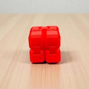 K&ouml;nnte beinhalten: Ein leuchtend rotes, w&uuml;rfelf&ouml;rmiges Fidget-Spielzeug. Das Spielzeug besteht aus mehreren verbundenen Bl&ouml;cken mit glatter, gl&auml;nzender Oberfl&auml;che. Es liegt auf einer hellen Holzmaserung, mit schlichtem wei&szlig;em Hintergrund.