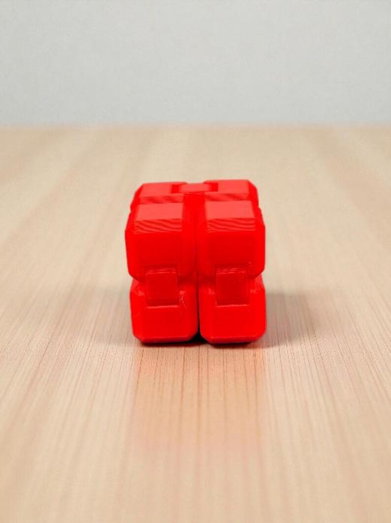 K&ouml;nnte beinhalten: Ein leuchtend rotes, w&uuml;rfelf&ouml;rmiges Fidget-Spielzeug. Das Spielzeug besteht aus mehreren verbundenen Bl&ouml;cken mit glatter, gl&auml;nzender Oberfl&auml;che. Es liegt auf einer hellen Holzmaserung, mit schlichtem wei&szlig;em Hintergrund.