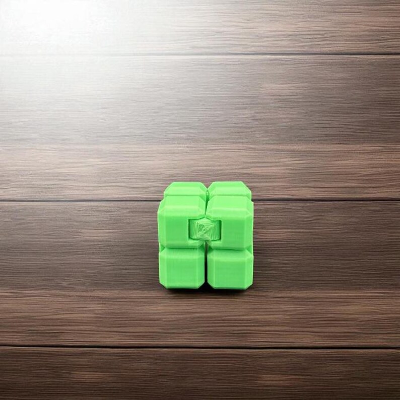 K&ouml;nnte beinhalten: Ein hellgr&uuml;ner Fidget-Cube, bestehend aus acht verbundenen Bl&ouml;cken. Der W&uuml;rfel besteht aus Kunststoff und steht auf einer braunen Holzoberfl&auml;che. Das Spielzeug ist zum Manipulieren und Spielen konzipiert.