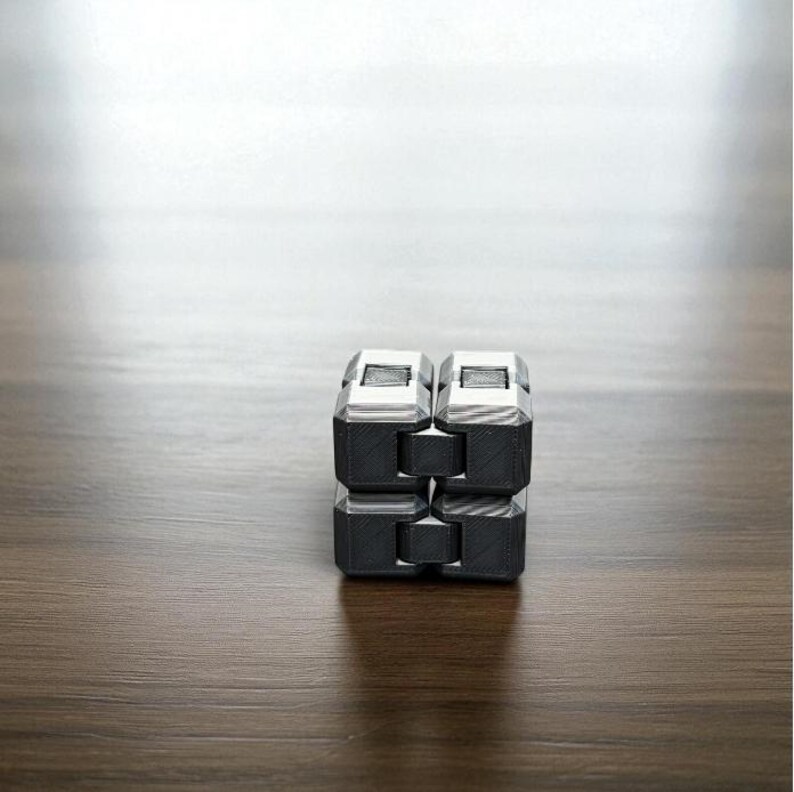 K&ouml;nnte beinhalten: Ein schwarzer und silberner Fidget Cube liegt auf einer Holzoberfl&auml;che. Der W&uuml;rfel besteht aus acht verbundenen Bl&ouml;cken, die gedreht und manipuliert werden k&ouml;nnen. Die oberen und unteren Bl&ouml;cke sind silberfarben, die mittleren Bl&ouml;cke sind schwarz.