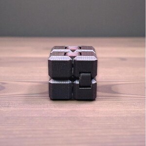 K&ouml;nnte beinhalten: Ein schwarzer Fidget Cube mit strukturierter Oberfl&auml;che. Der W&uuml;rfel besteht aus mehreren verbundenen Bl&ouml;cken, die manipuliert werden k&ouml;nnen. Der W&uuml;rfel liegt auf einer Holzoberfl&auml;che.