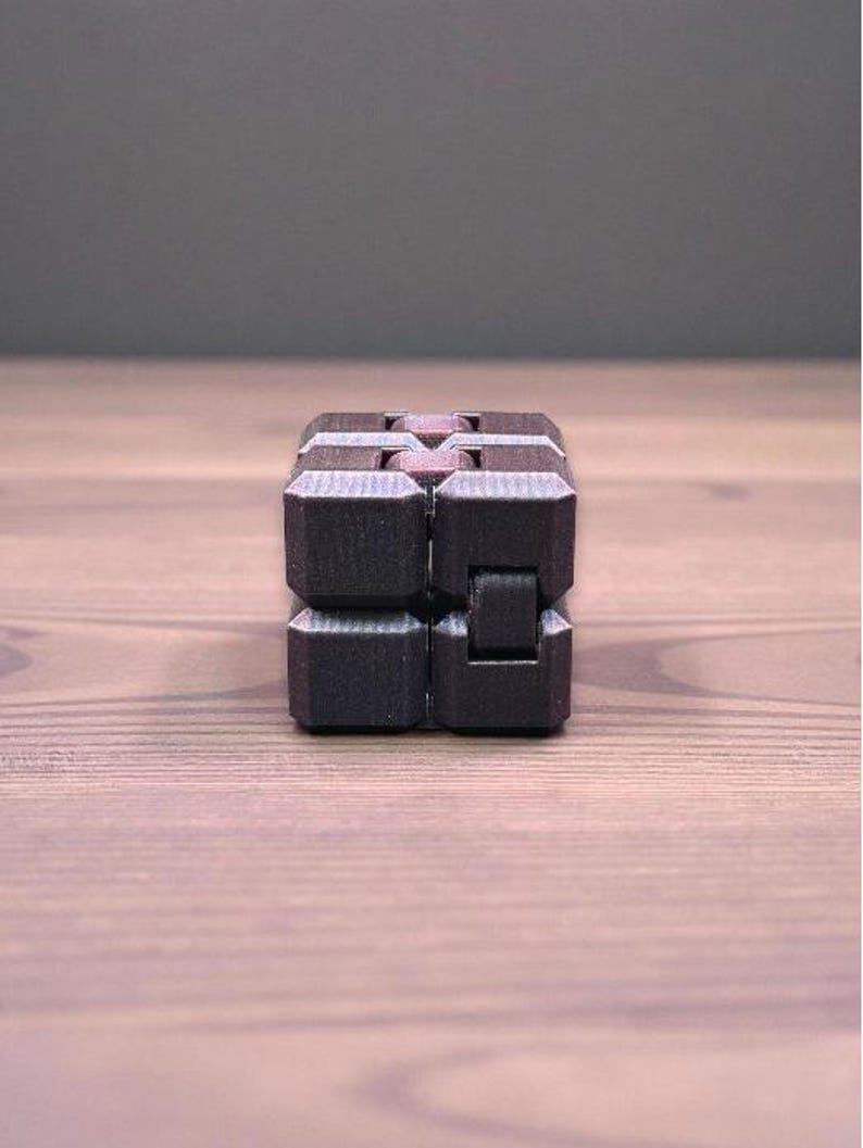 K&ouml;nnte beinhalten: Ein schwarzer Fidget Cube mit strukturierter Oberfl&auml;che. Der W&uuml;rfel besteht aus mehreren verbundenen Bl&ouml;cken, die manipuliert werden k&ouml;nnen. Der W&uuml;rfel liegt auf einer Holzoberfl&auml;che.