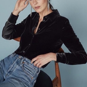 Op de afbeelding: Een zwart fluwelen overhemd met lange mouwen en een kraag, gecombineerd met lichtblauwe jeans. Het model zit op een houten stoel tegen een lichtblauwe achtergrond. Het model draagt lange zilveren oorbellen.