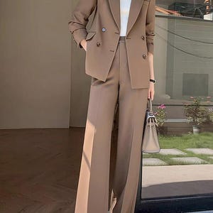 Pode incluir: Um fato de neg&oacute;cios castanho com um blazer de abotoamento duplo e cal&ccedil;as de perna larga. O fato &eacute; combinado com uma gola alta branca e uma mala de m&atilde;o de cor clara. O conjunto &eacute; de cor neutra, adequado para ambientes profissionais.