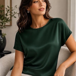 May include: A dark forest green short-sleeved blouse with a smooth, satin-like finish and a round neckline. The garment features a relaxed fit with cuffed sleeves and a subtle sheen that reflects light across the fabric surface.