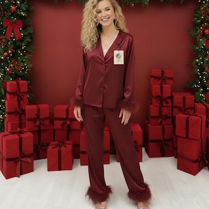 May include: Burgundy satin pyjama set with feather trim on the cuffs and ankles. The set includes a long-sleeved button-down shirt and matching trousers. The model is wearing gold sandals. The background features red gift boxes and Christmas decorations.