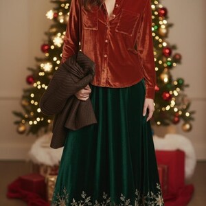 May include: A rust-coloured velvet button-down shirt with two chest pockets and a long-sleeved design. Paired with a long, emerald green velvet skirt with gold floral embroidery at the hem. A brown jacket is held in the arm.