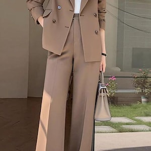 Pode incluir: Um fato castanho composto por um blazer de abotoamento duplo e cal&ccedil;as de perna larga. O blazer tem duas filas de bot&otilde;es e o conjunto &eacute; combinado com um top branco. Uma mala clara &eacute; segurada na m&atilde;o.