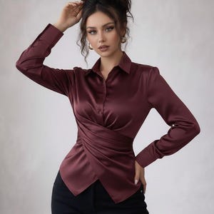 Könnte beinhalten: Eine bordeauxfarbene Satinbluse mit Wickeldesign. Das langärmlige Hemd hat einen Kragen und ist vorne geknöpft. Die Bluse wird mit einer schwarzen Hose kombiniert. Das Model trägt goldene Ohrringe.