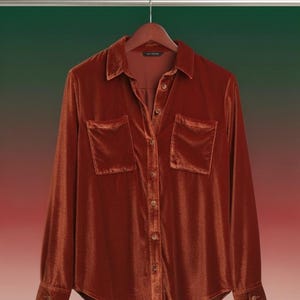 May include: A burnt orange velvet shirt with long sleeves, a collared neckline, and two front pockets. The shirt is button-down and hangs on a wooden hanger against a green and red gradient background.